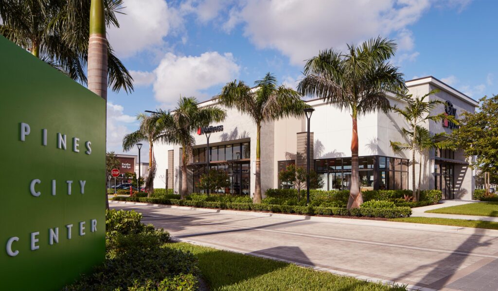 Landscaped entrance to Pines City Center with tall palm trees, flower beds, brick walkways, and storefront signs along Pines Boulevard in Pembroke Pines, Florida