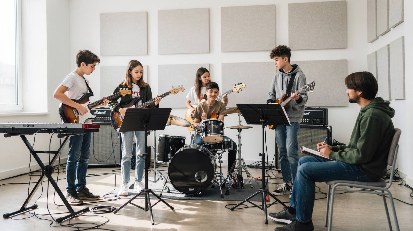 Youth band practice session with teenagers playing electric guitar, bass, drums and keyboard in a bright rehearsal room