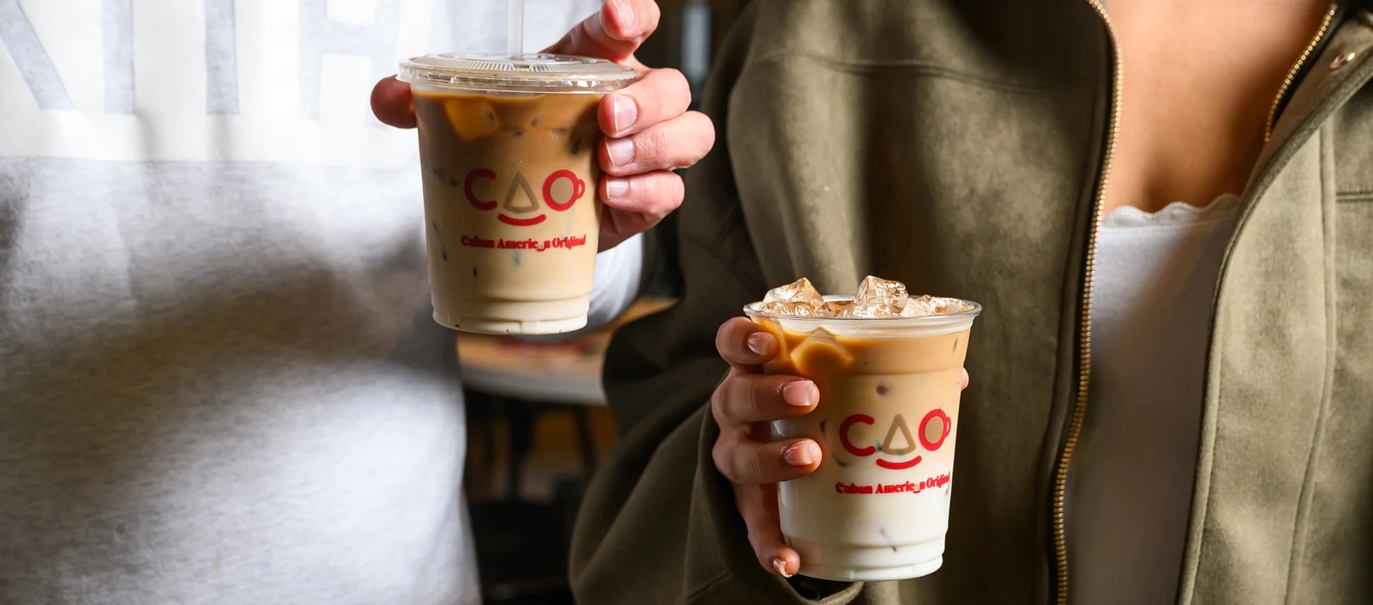 Two people toast CAO iced Cuban coffees in clear plastic cups with the red 'CAO Cuban American Original' smiley logo — one in a grey sweatshirt, one in an olive jacket