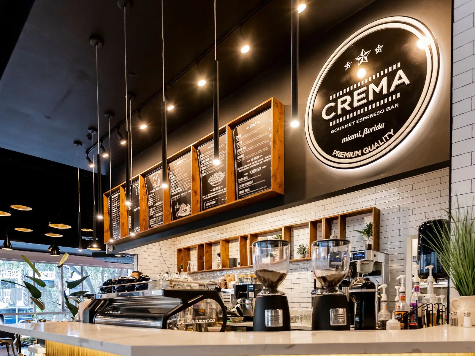Crema Gourmet Espresso Bar Pembroke Pines — illuminated CREMA sign above the coffee counter with espresso machine, grinders and hanging menu boards