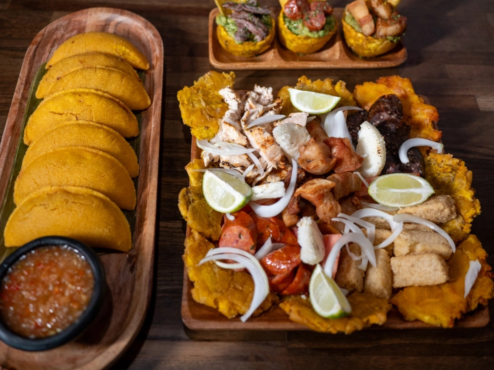 El Machetico Colombian spread — golden empanadas with salsa on a wooden tray beside a large platter of tostones topped with shredded chicken, sausage and pork with lime wedges, and three plantain cups of guacamole topped with beef, chorizo and pork belly