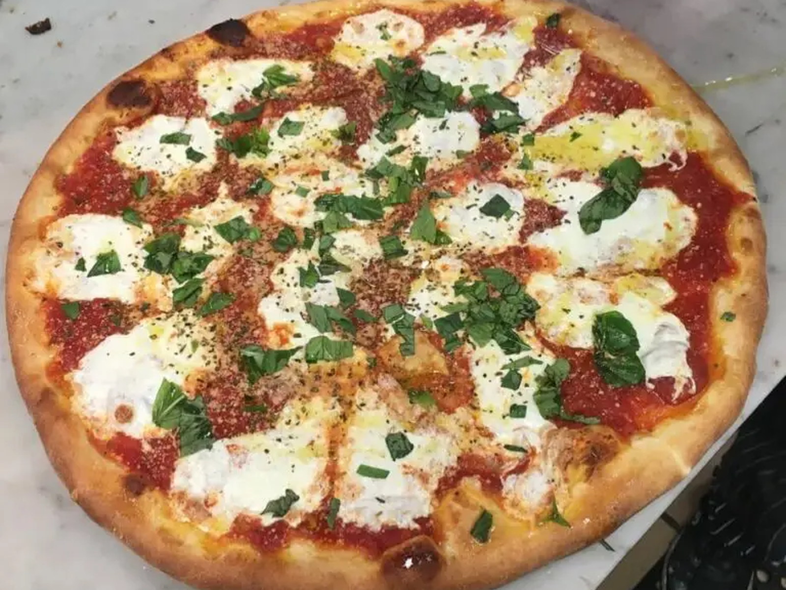 Pasquale's Pizza Company Margherita-style pizza on a white marble counter — golden, slightly charred crust, vibrant tomato sauce, dollops of melted fresh mozzarella, chopped basil, a sprinkle of dried herbs and a drizzle of olive oil